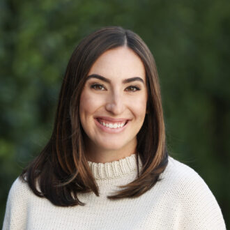 A woman with straight brown hair wearing a cream-colored sweater with black stripes smiles at the camera, standing outdoors with a blurred green background.
