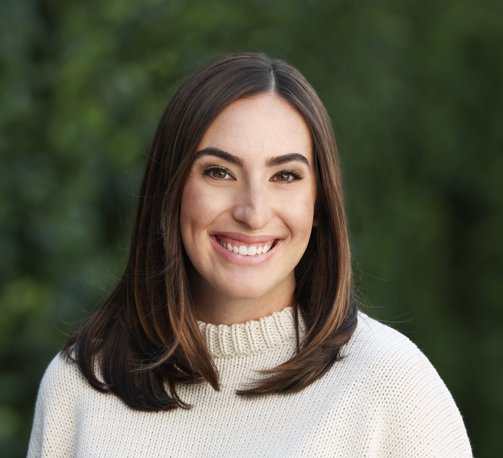 A woman with straight brown hair wearing a cream-colored sweater with black stripes smiles at the camera, standing outdoors with a blurred green background.