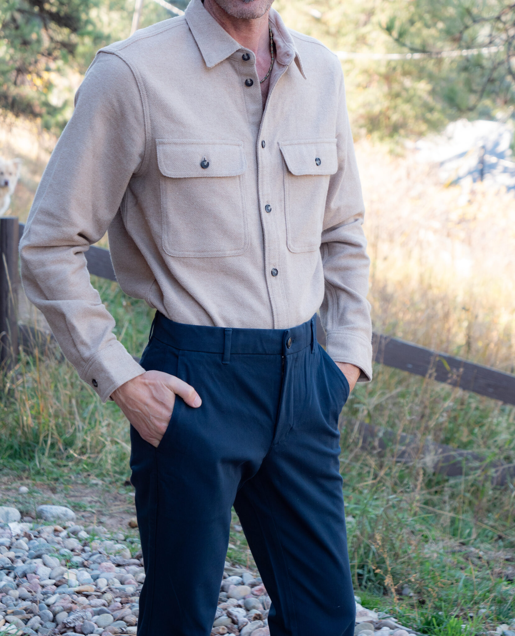 A person stands outdoors wearing a beige button-up shirt with chest pockets and dark blue trousers, with hands in pockets.