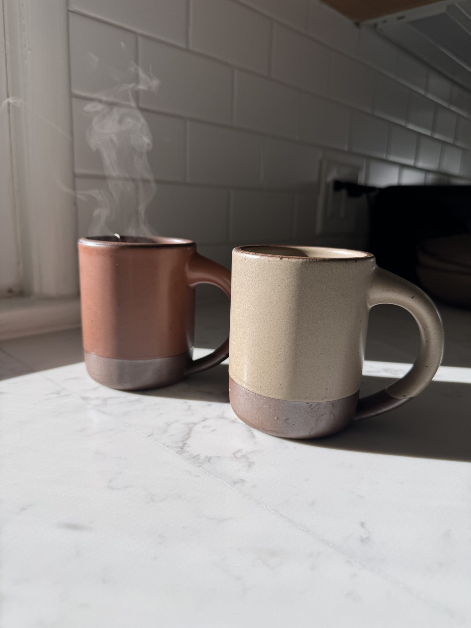Two ceramic mugs, one brown and one beige, sit on a sunlit marble countertop; steam rises from the brown mug. White subway tile backsplash in the background.