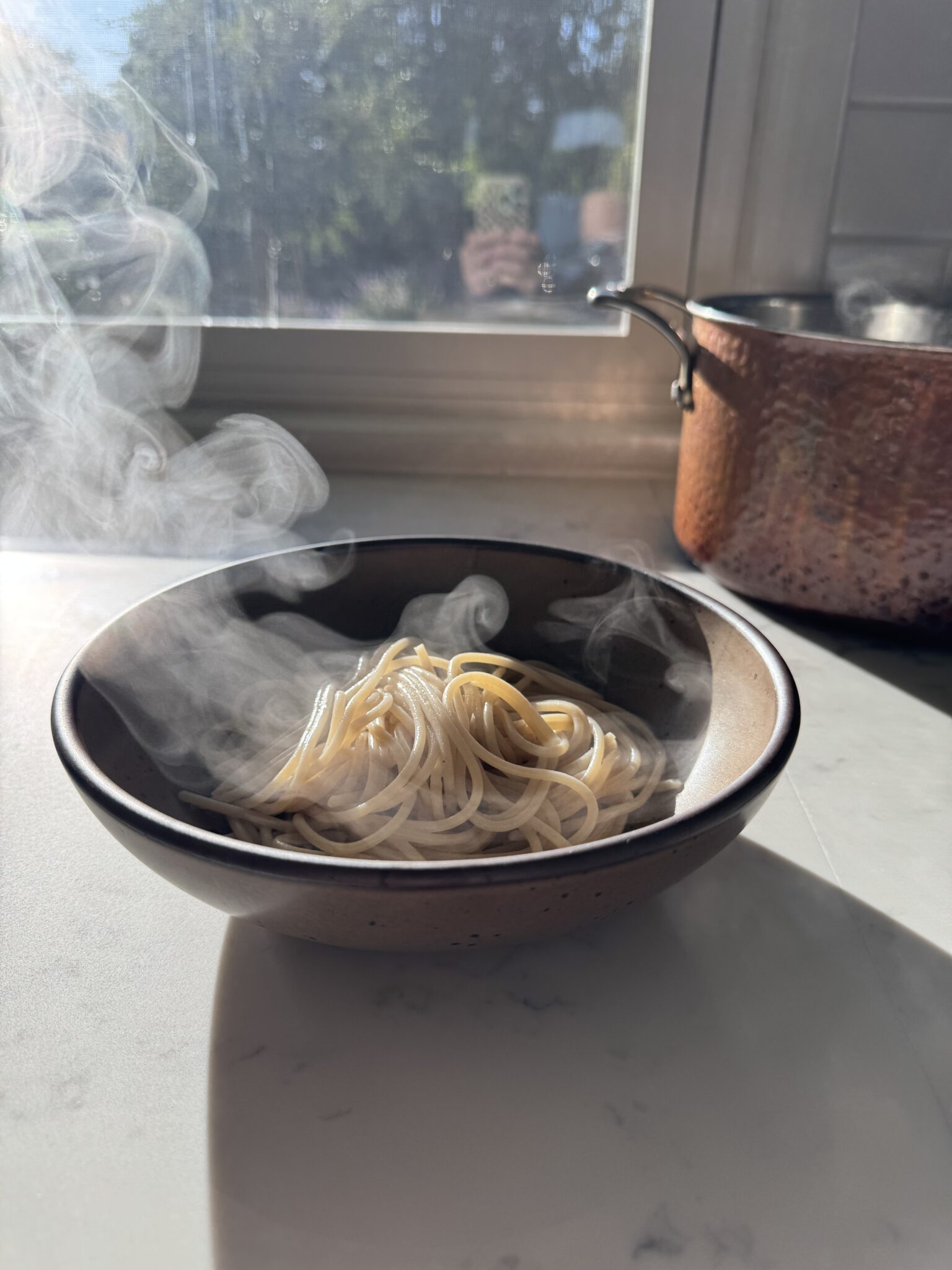 A bowl of steaming cooked spaghetti sits on a sunlit countertop near a window, with a copper pot in the background.