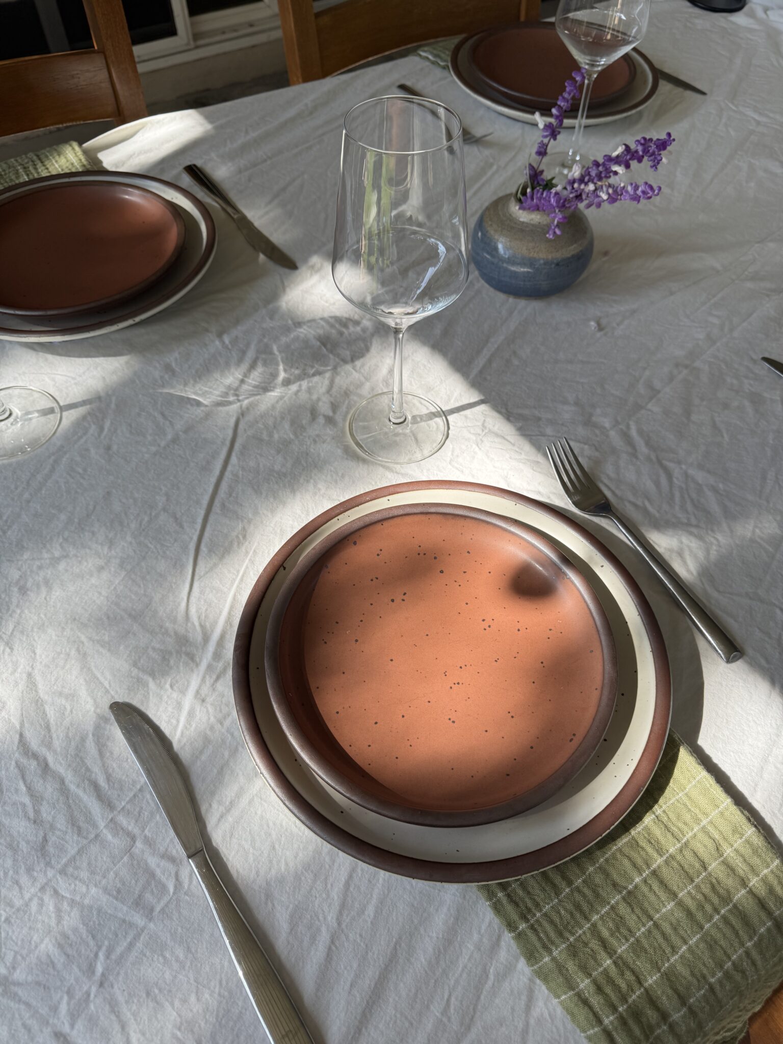 A set dining table with ceramic plates, a wine glass, cutlery, a green striped napkin, and a small vase with purple flowers on a white tablecloth.