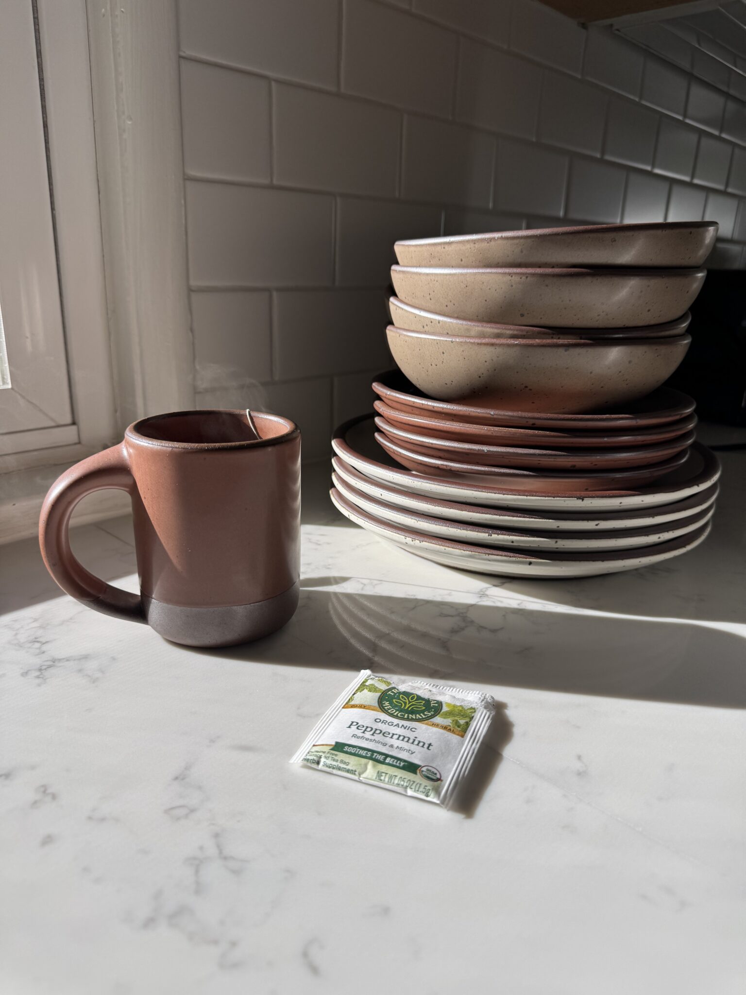 A brown ceramic mug, a stack of pink and white bowls and plates, and a packet of peppermint tea sit on a marble countertop in sunlight.