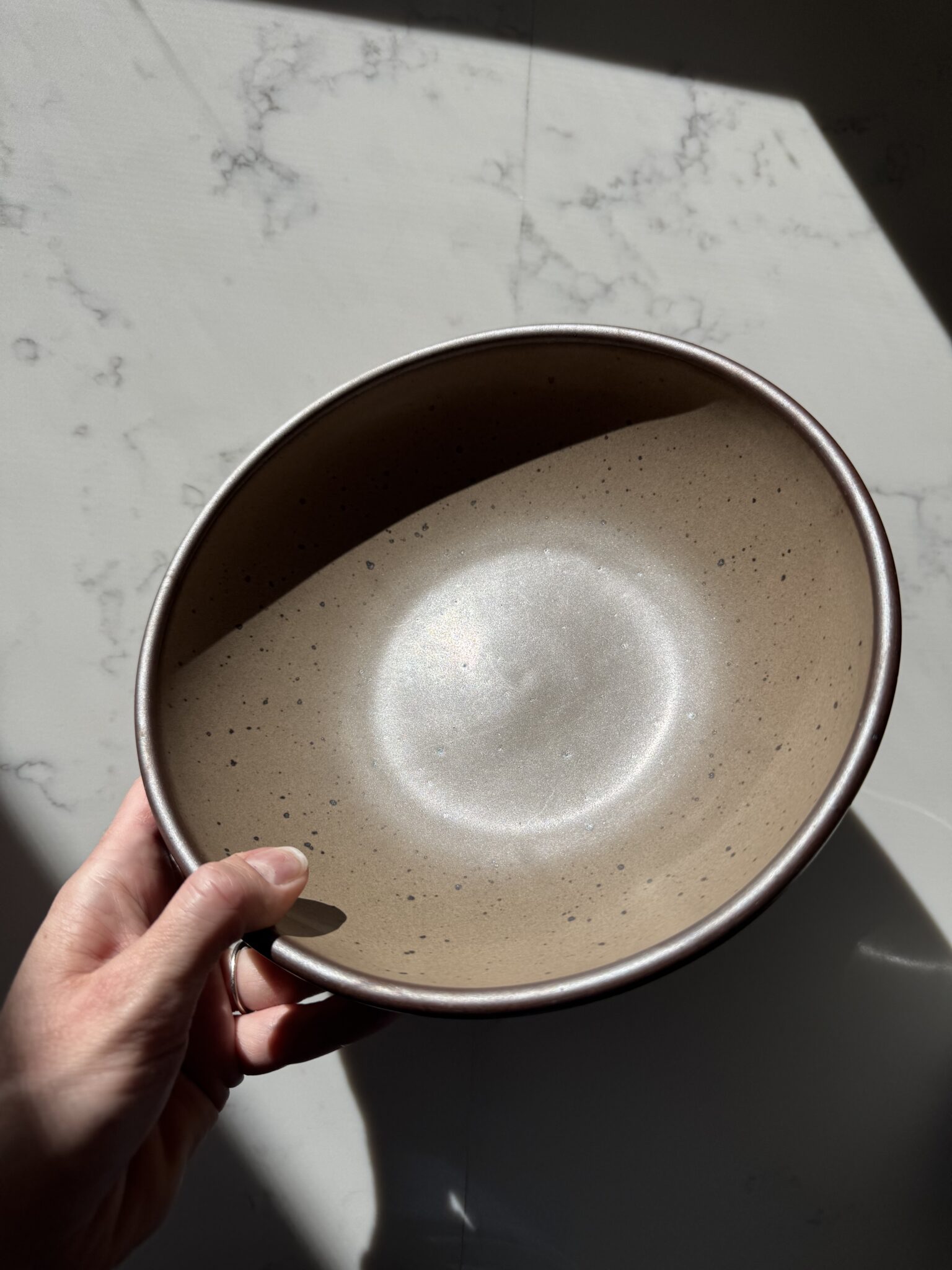 A hand holds a round, beige ceramic bowl with a speckled pattern, positioned under natural sunlight on a marbled surface.