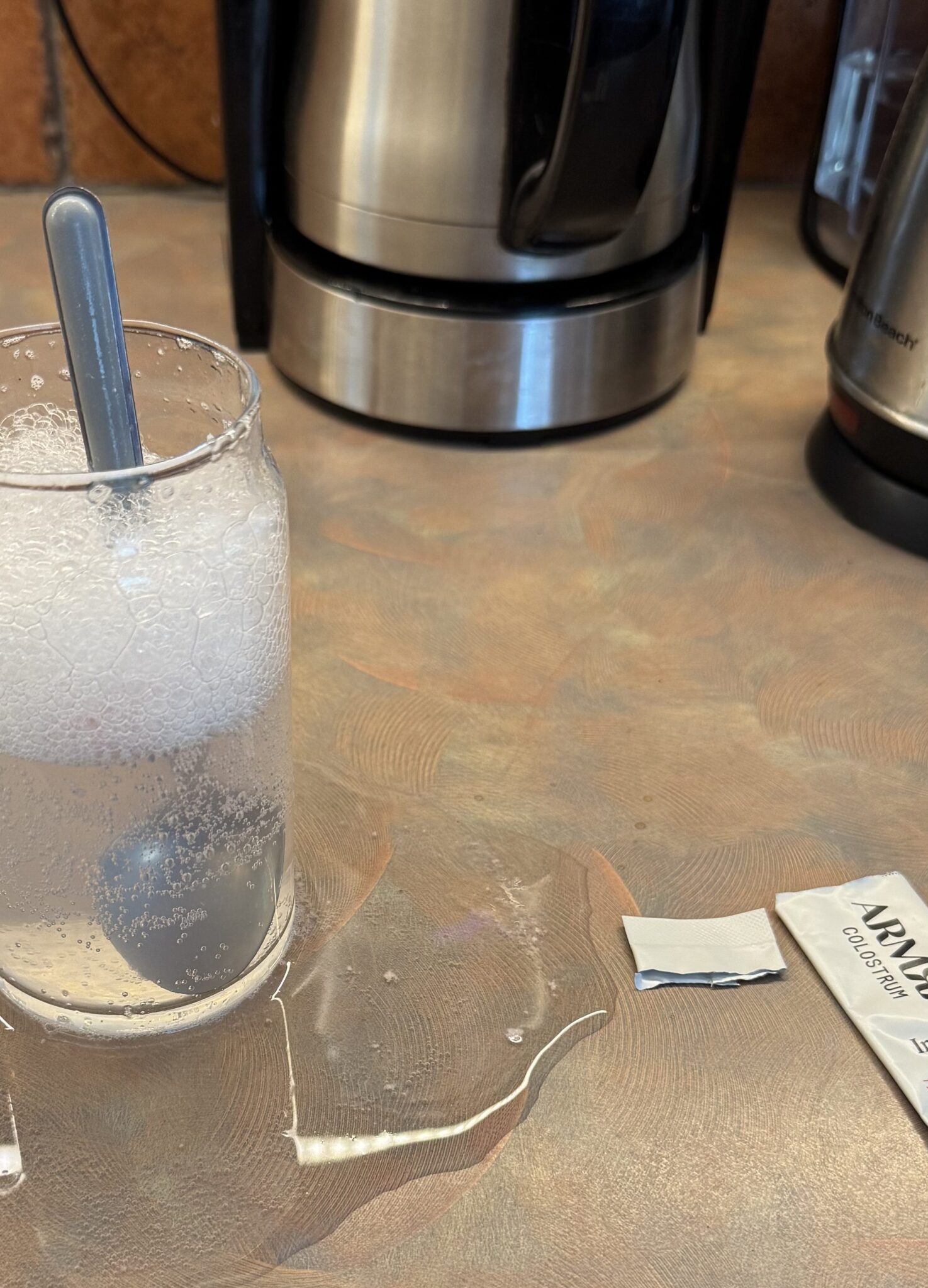 A glass of fizzy liquid with a spoon sits on a kitchen counter, surrounded by spilled liquid and an empty Lypo-Spheric Vitamin C packet.