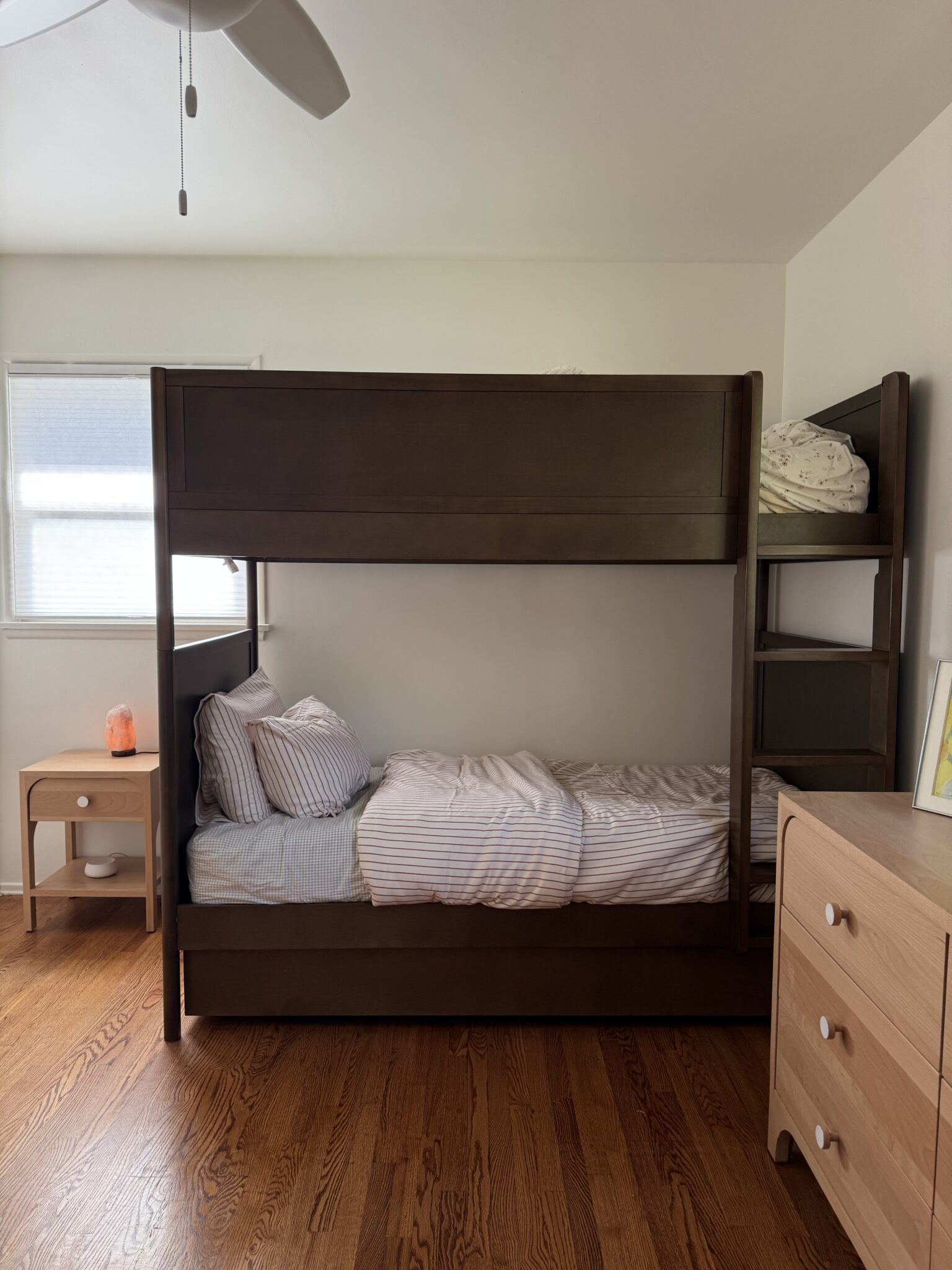 A wooden bunk bed with striped bedding sits against a wall in a tidy bedroom with hardwood floors, a nightstand, dresser, and a ceiling fan visible.
