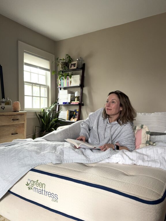 A woman sits up in bed reading a book, with a bookshelf and window in the background. The bed has a "my green mattress" label visible on the side.