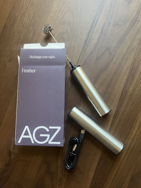 A stainless steel handheld milk frother with a whisk, USB charging cable, and purple AGZ-branded box are displayed on a wooden surface.