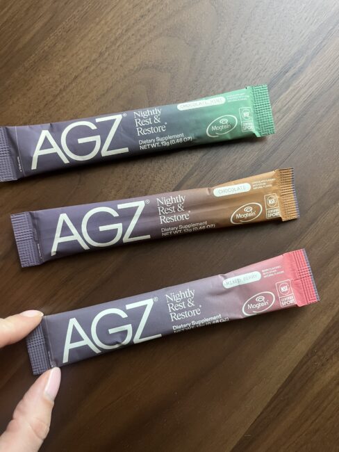 Three AGZ Nightly Rest & Restore dietary supplement packets in chocolate mint, chocolate, and mixed berry flavors are displayed on a wooden surface. A hand holds the mixed berry packet.
