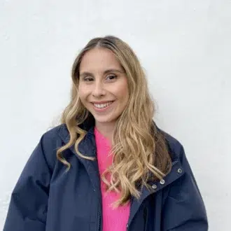 A person with long blonde hair, wearing a dark jacket over a bright pink top, stands in front of a plain white wall and smiles at the camera.