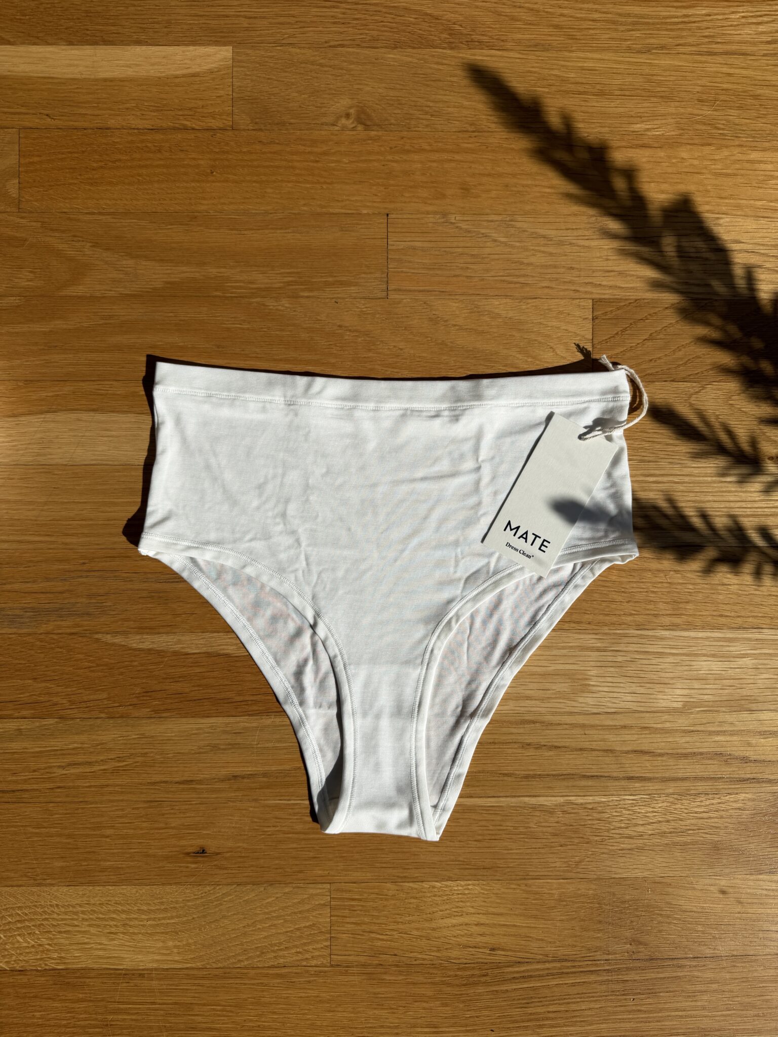 White high-waisted underwear with a tag labeled "MATE" displayed on a wooden surface, with a shadow of a plant on the right side.