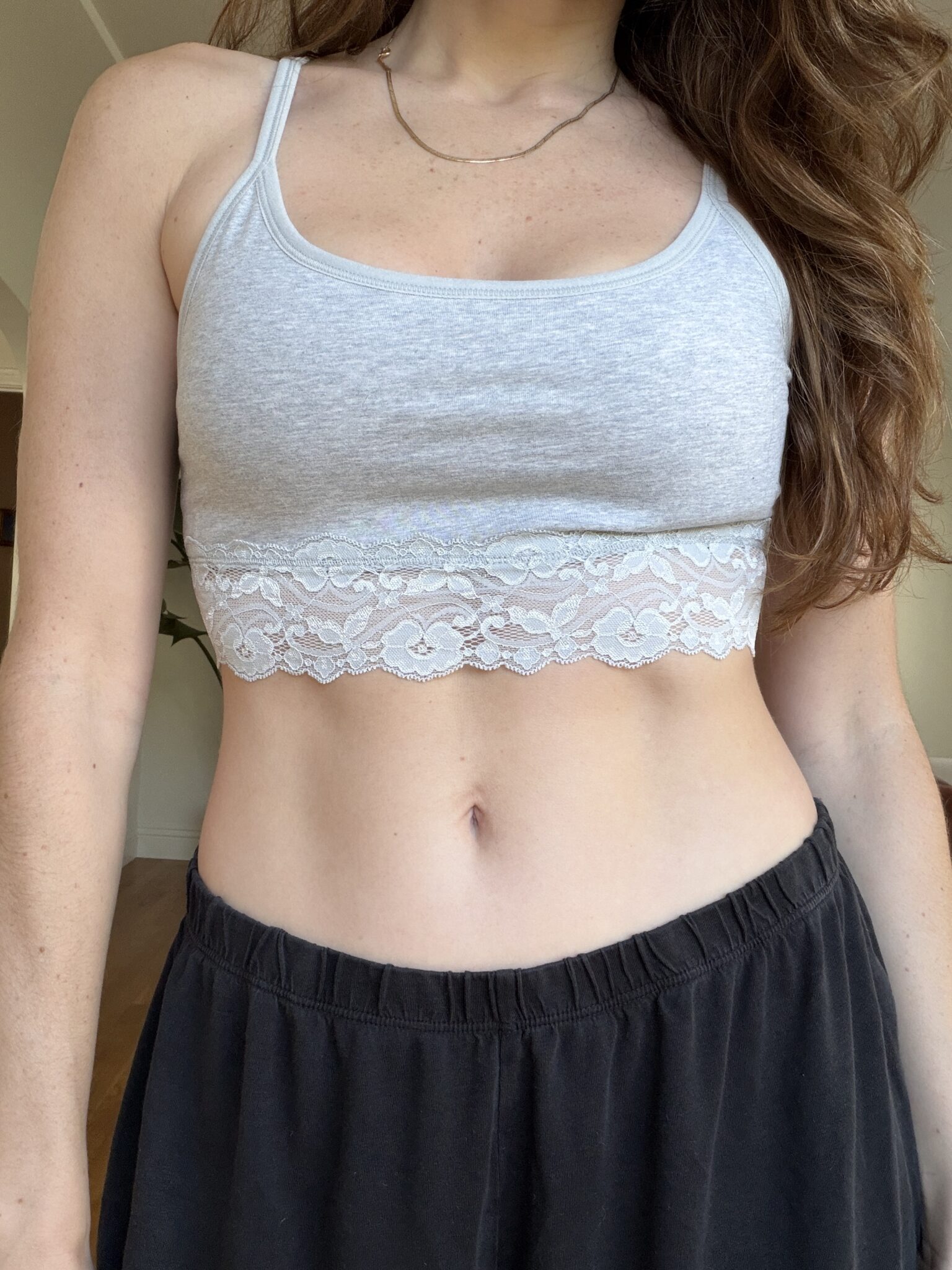 A person wearing a light gray lace-trimmed bralette and black pants stands indoors, shown from shoulders to waist.