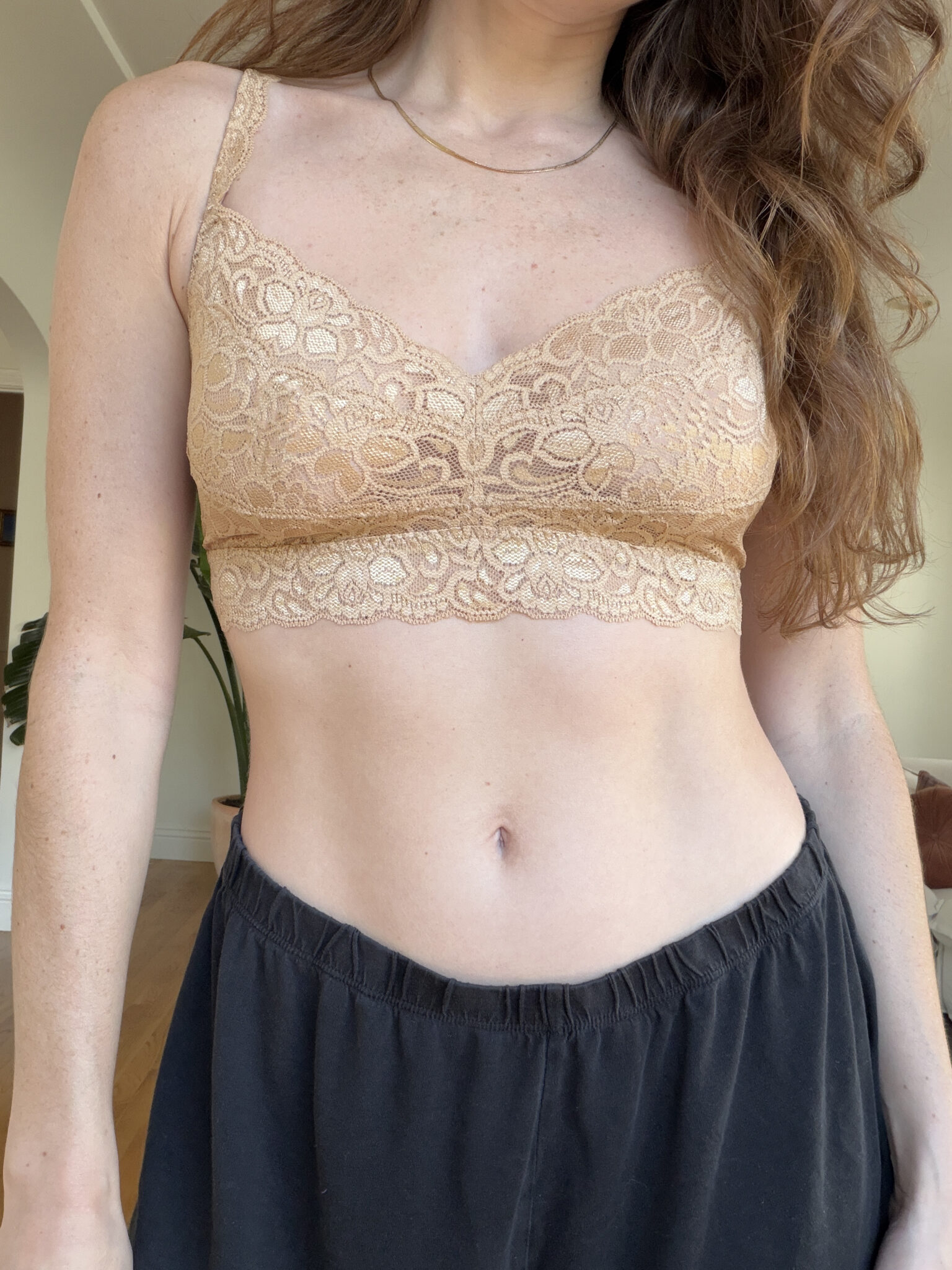 A person wearing a beige lace bralette and black pants, standing indoors with long hair partially visible.