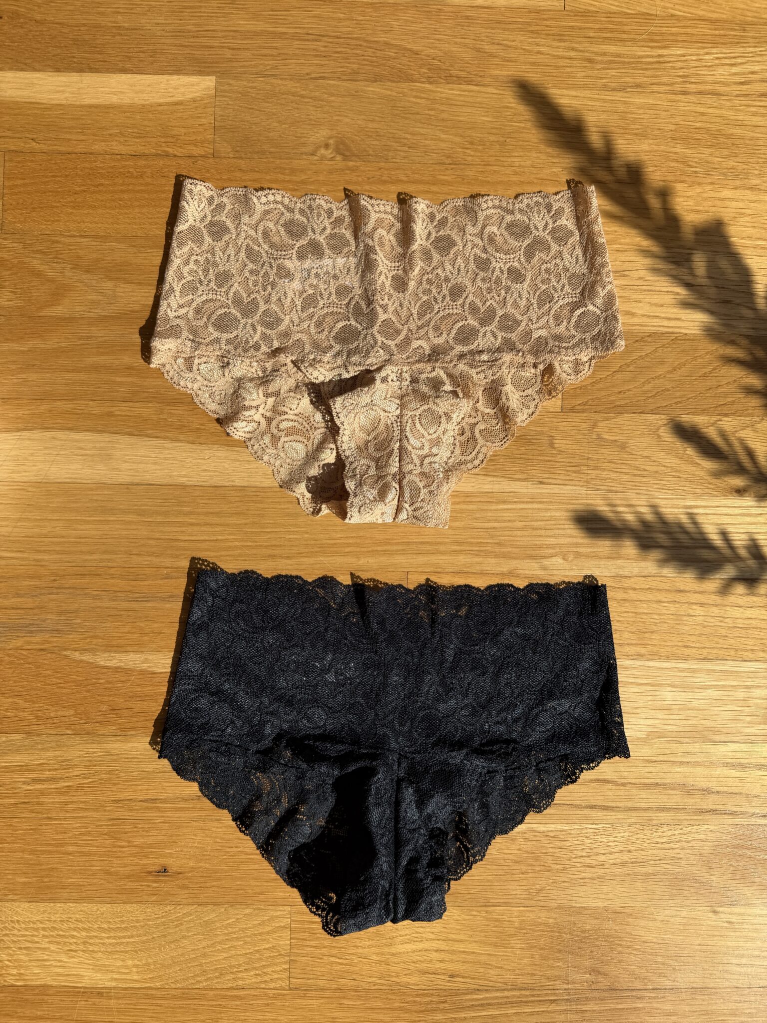 Two pairs of lace underwear, one beige and one black, are laid flat on a wooden surface in sunlight.
