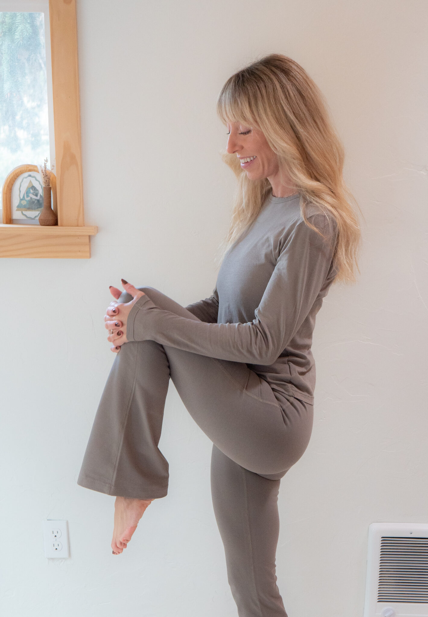 A woman in gray loungewear stands indoors, lifting one knee to her chest while smiling. She is next to a window with light coming through.