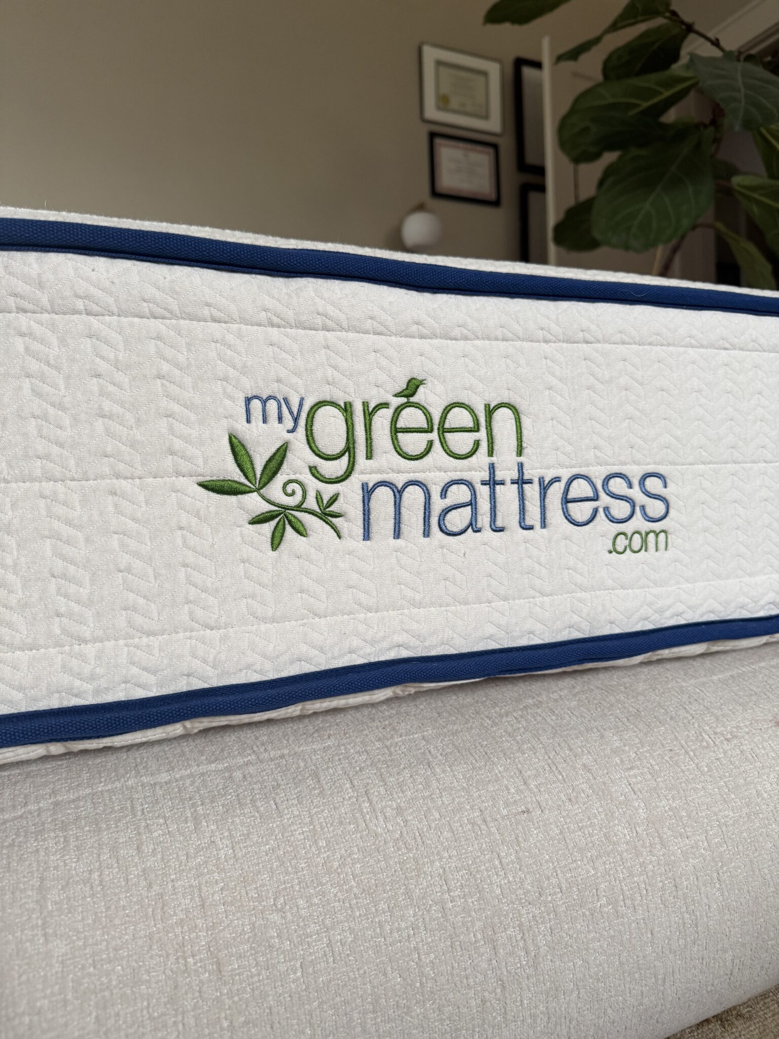 Close-up of a mattress side panel displaying the embroidered logo "mygreenmattress.com" with leaf graphics, against a blurred indoor background with certificates and a plant.