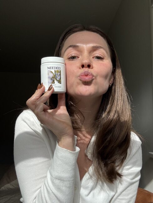 A woman wearing a white top holds up a container labeled "NEEDED" and poses with puckered lips in natural light.