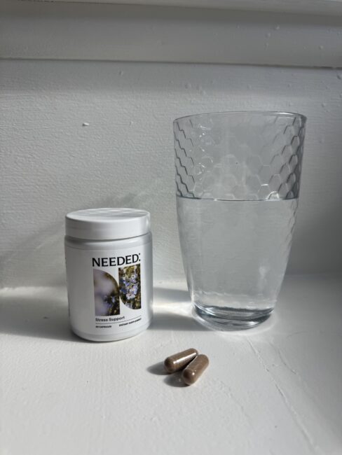 A container labeled "Needed: Stress Support," two brown capsules, and a glass of water are placed on a white surface.