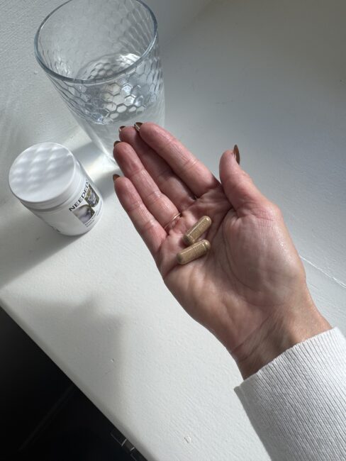 A hand holding two brown capsules above a white surface, with a glass of water and a white supplement container nearby.