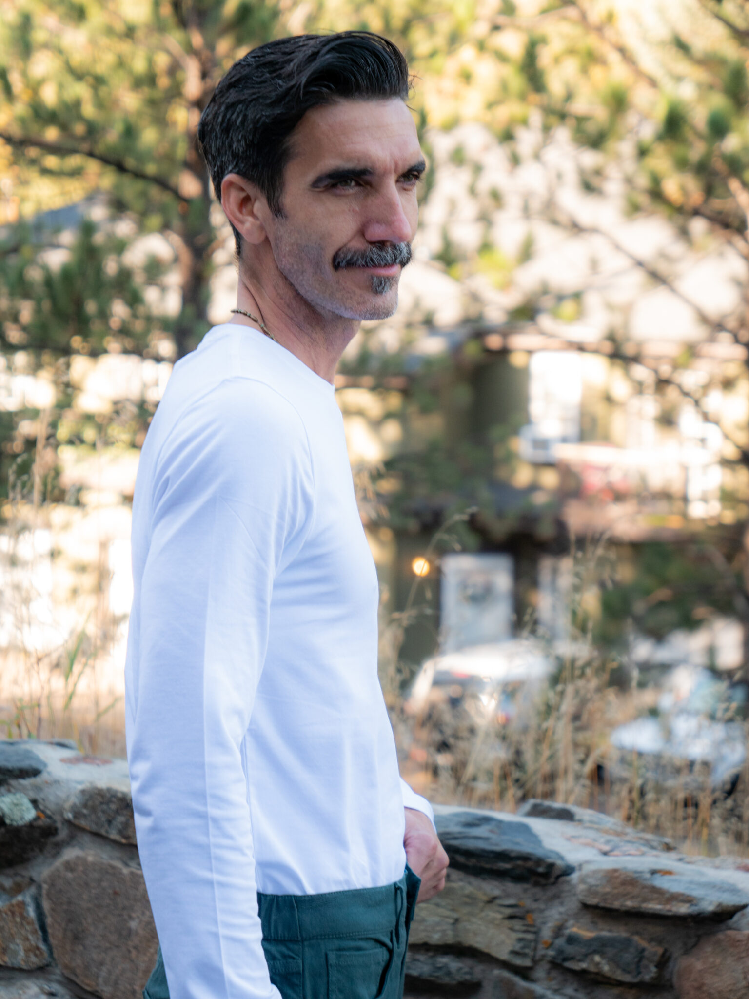 A man with dark hair and a mustache, wearing a white long-sleeve shirt and green pants, stands outdoors near a stone wall with trees and buildings in the blurred background.