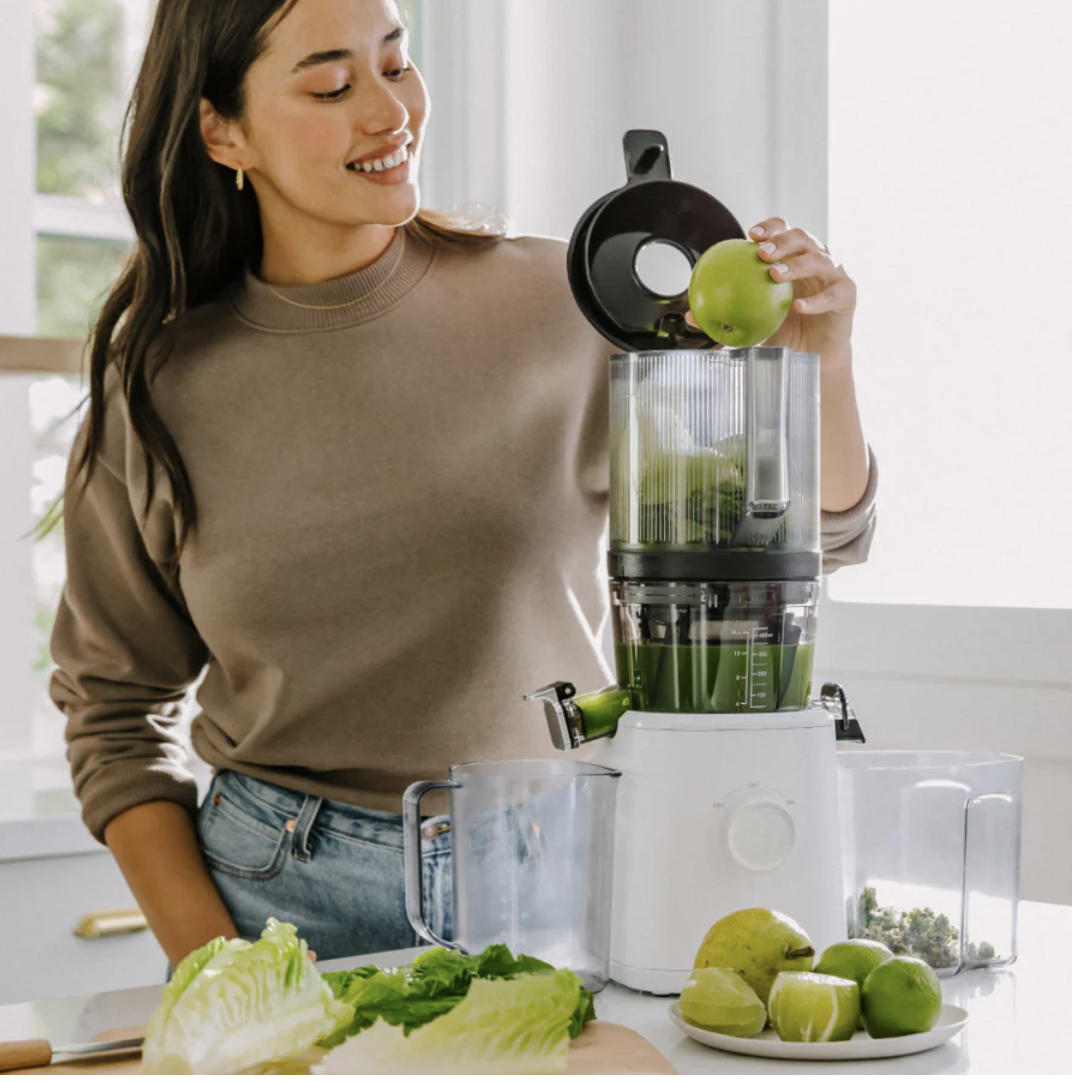 A woman places a green apple into a juicer on a kitchen counter, surrounded by apples, lettuce, and juice containers.