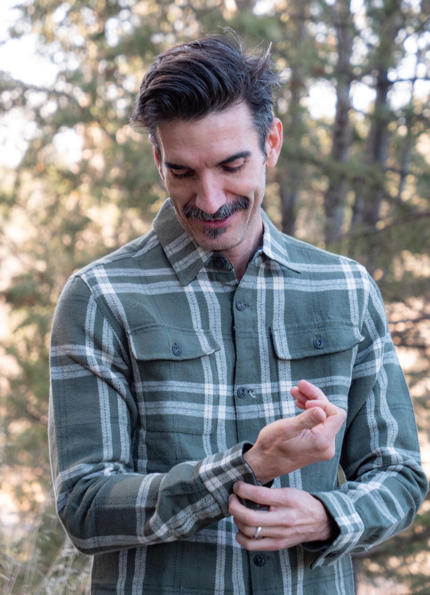 A man with a mustache wearing a green plaid shirt stands outdoors, looking down and adjusting his sleeve. Trees are visible in the background.