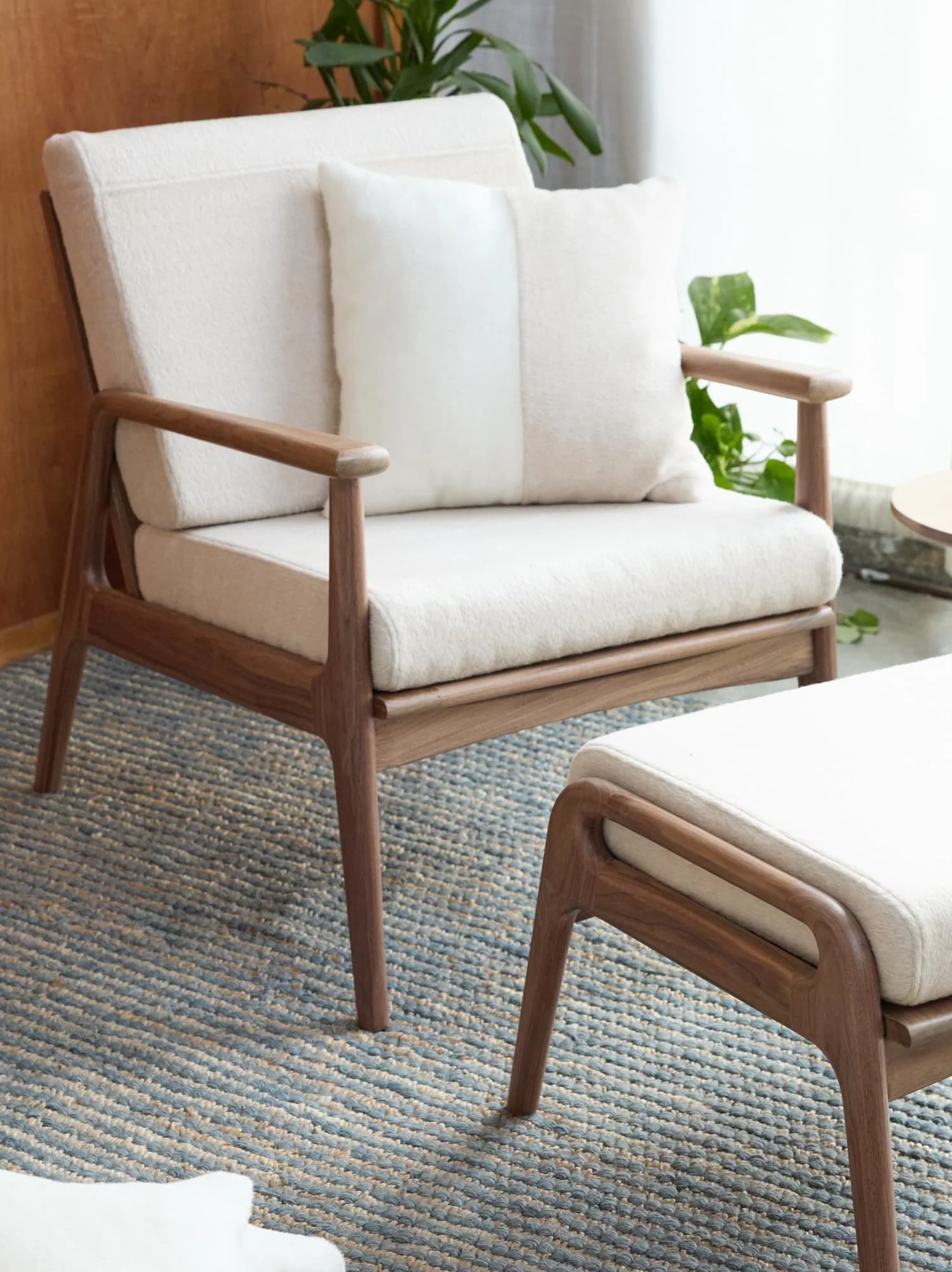 A cushioned wooden armchair from sustainable brands with a white pillow sits on a textured blue-gray rug next to a matching footrest, with plants and a window in the background.