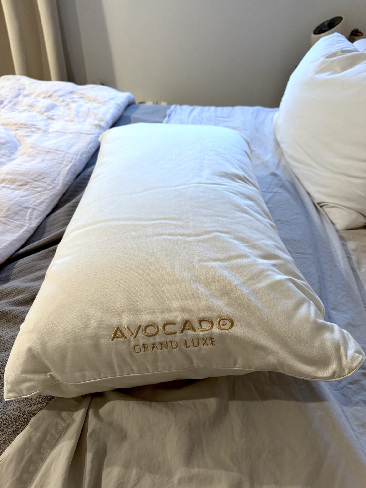 A white pillow with "Avocado Grand Luxe" embroidered in gold rests on a neatly made bed with white bedding.
