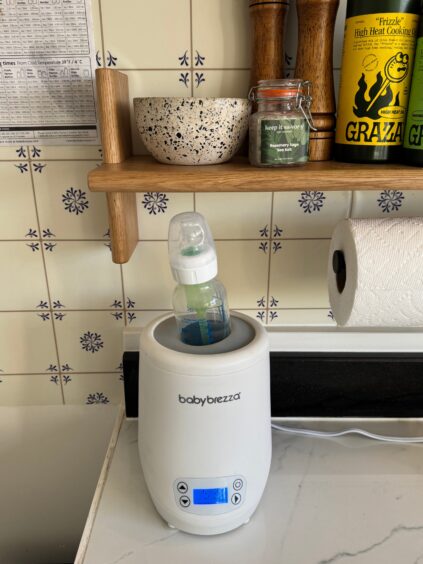 A baby bottle is being warmed in a Baby Brezza bottle warmer on a kitchen counter, with shelves and various kitchen items in the background.