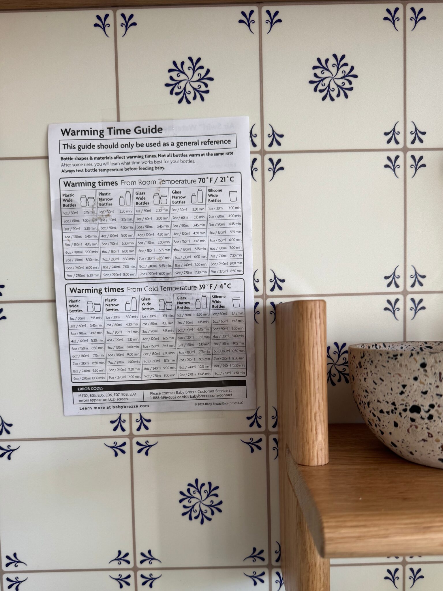 A printed warming time guide is taped to a kitchen wall with decorative white tiles, next to a wooden shelf holding a ceramic bowl.