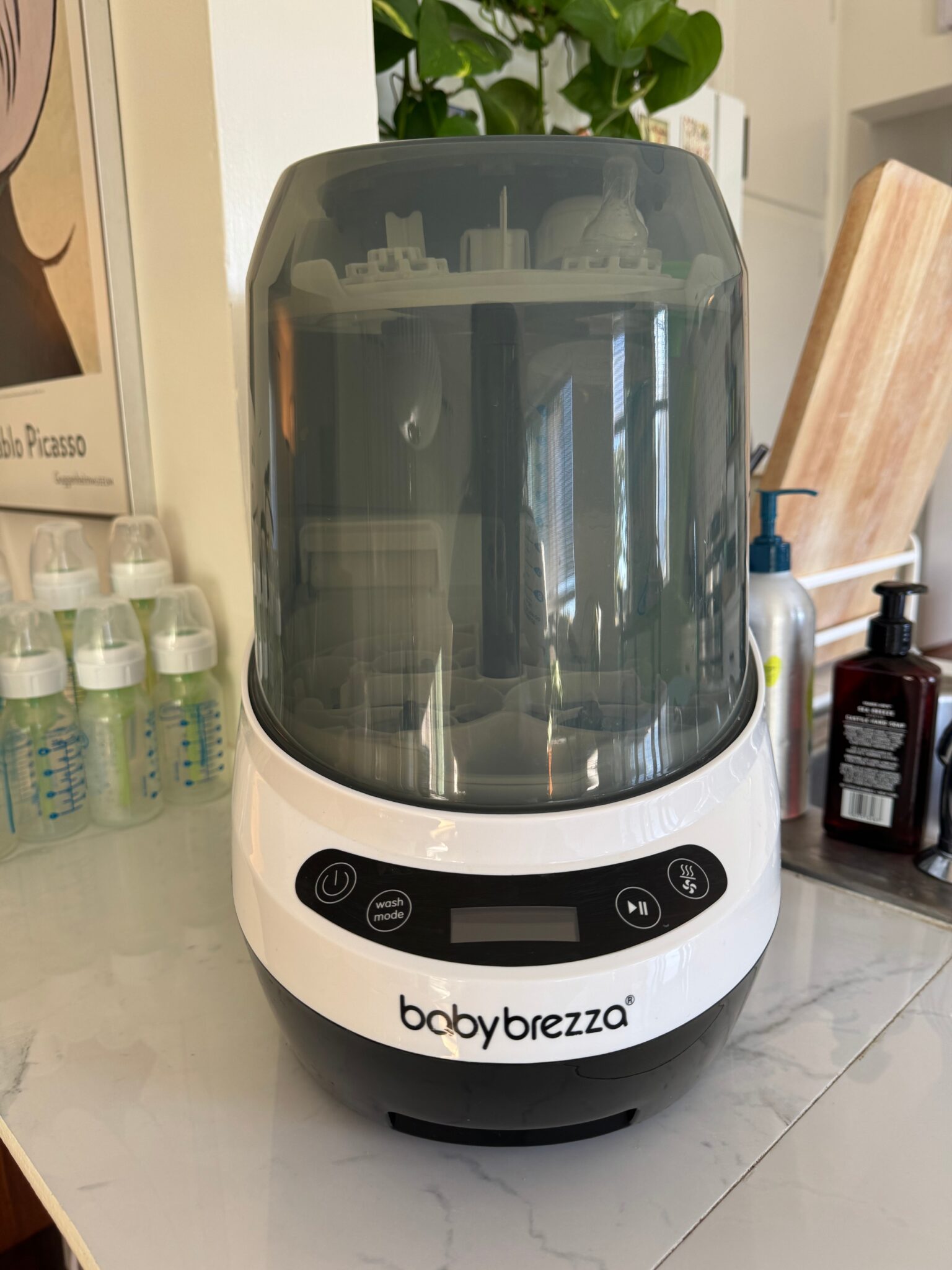 A Baby Brezza sterilizer and dryer sits on a kitchen counter, with baby bottles, soap dispensers, and a cutting board in the background.