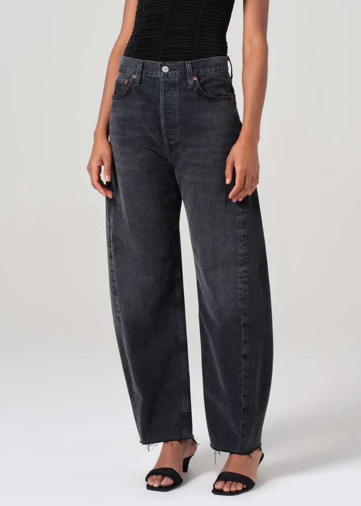 Person wearing wide-leg, high-waisted black jeans with a frayed hem, a black sleeveless top, and black open-toe heels, standing against a plain background.