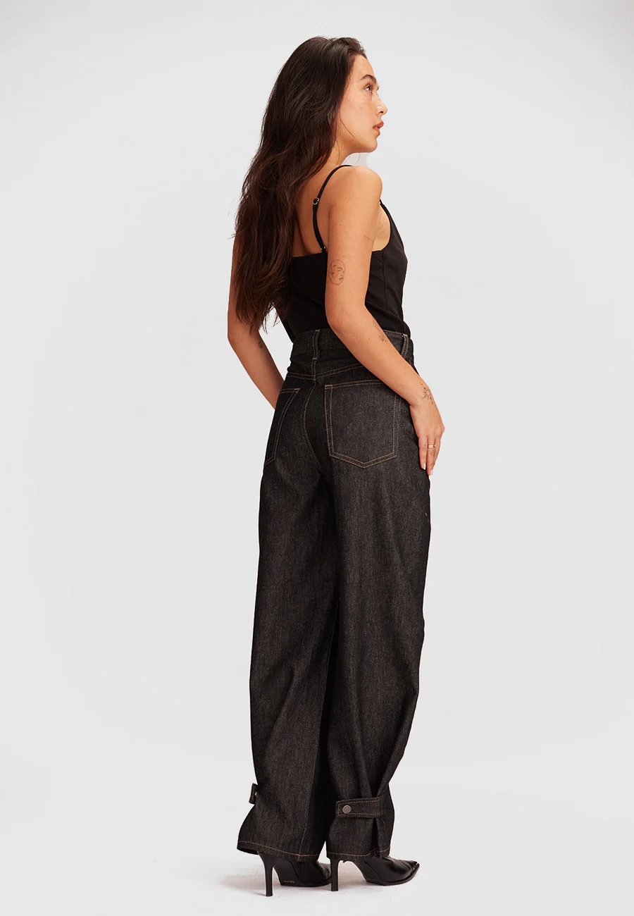 Woman standing in profile wearing a black tank top, high-waisted wide-leg dark jeans, and black heeled shoes against a plain light background.