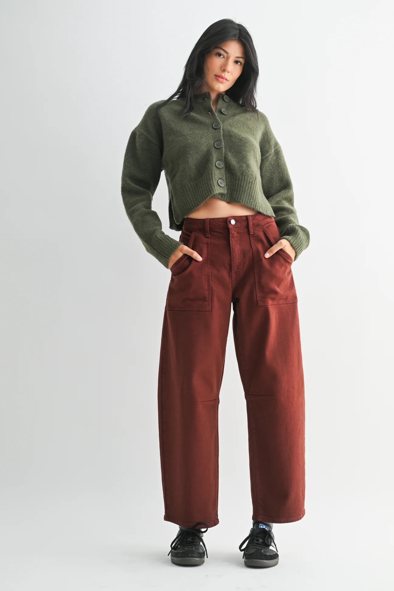Woman standing against a plain background, wearing an olive green button-up sweater, rust-colored wide-leg pants, and black sneakers, with hands in her pockets.