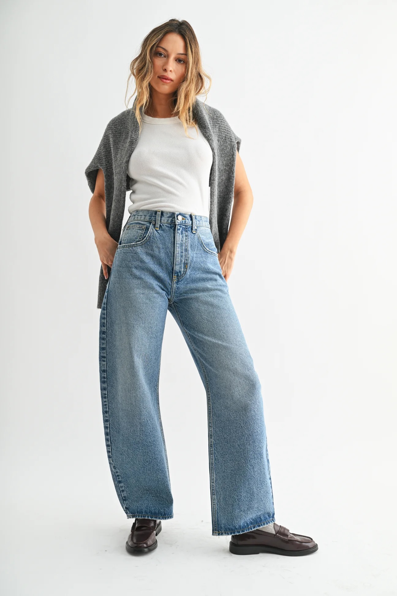 Woman standing against a plain background, wearing a grey sweater draped over shoulders, a white sleeveless top, high-waisted wide-leg blue jeans, and dark loafers.