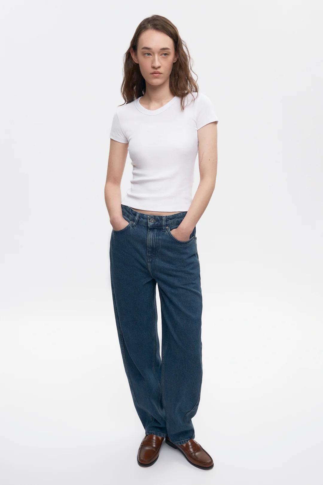 A person stands against a plain white background wearing a white short-sleeve t-shirt, loose blue jeans, and brown shoes, with hands in pockets.