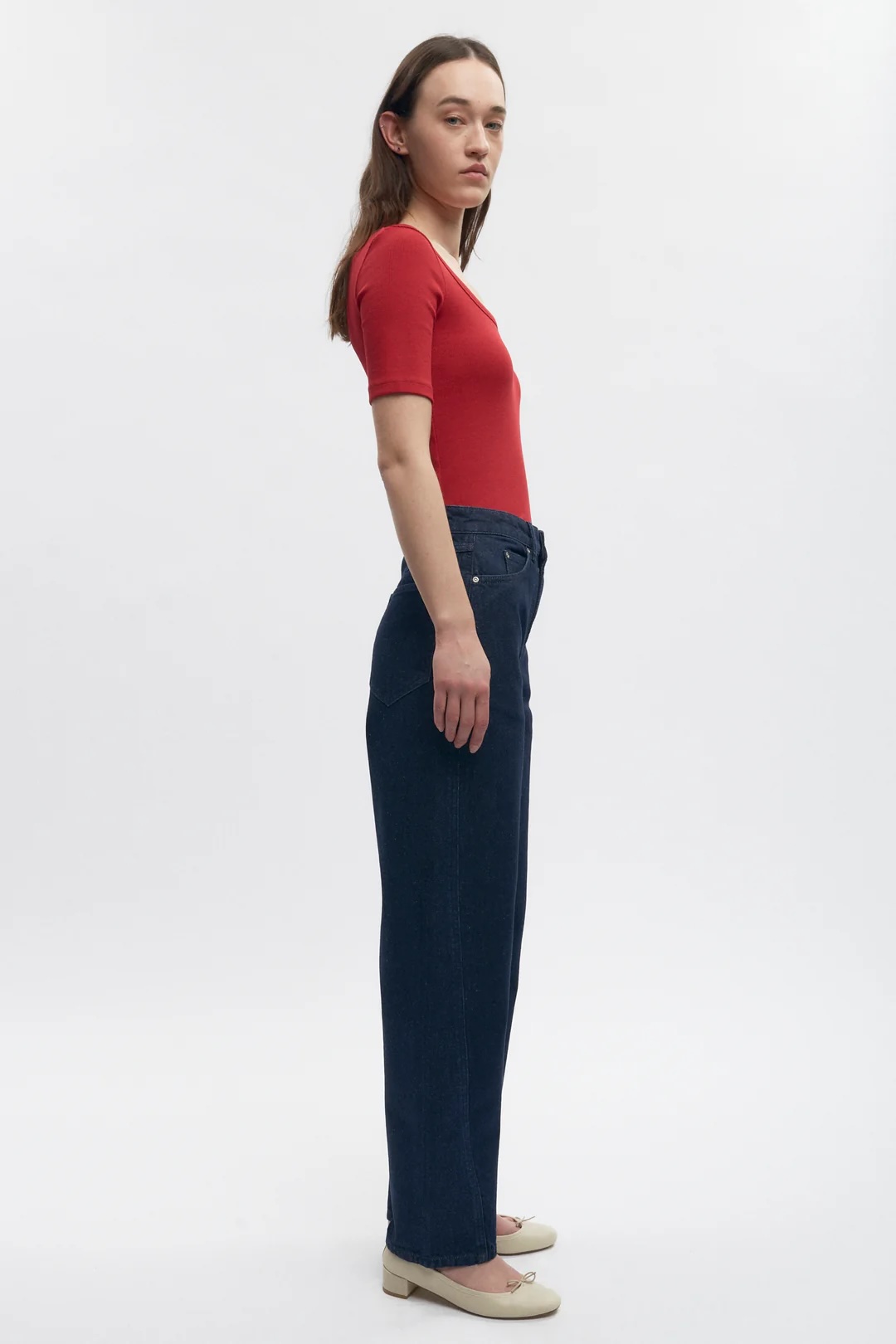 A woman stands in profile against a white background, wearing a red short-sleeve top, dark blue jeans, and cream-colored shoes.