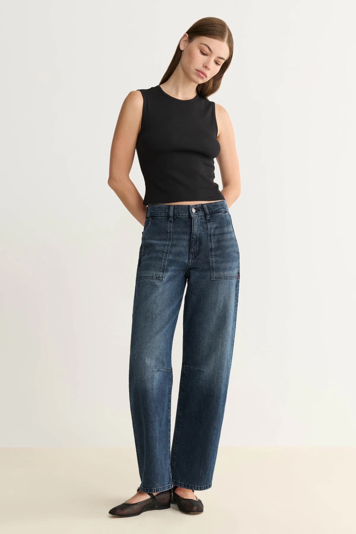 Woman standing against a plain background, wearing a sleeveless black top, high-waisted dark blue jeans, and black flat shoes, with hands behind her back.