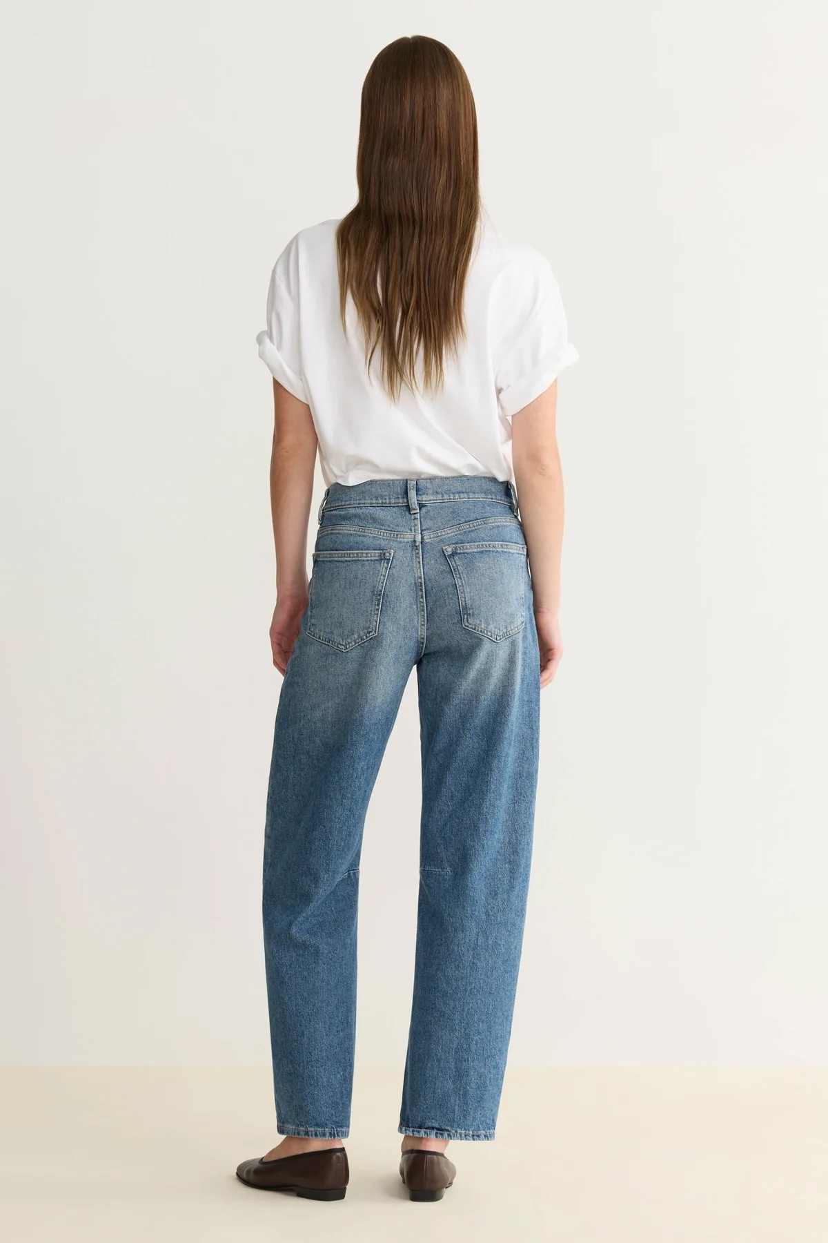 Person standing with back facing the camera, wearing a white short-sleeve shirt, blue high-waisted jeans, and brown flat shoes against a plain background.