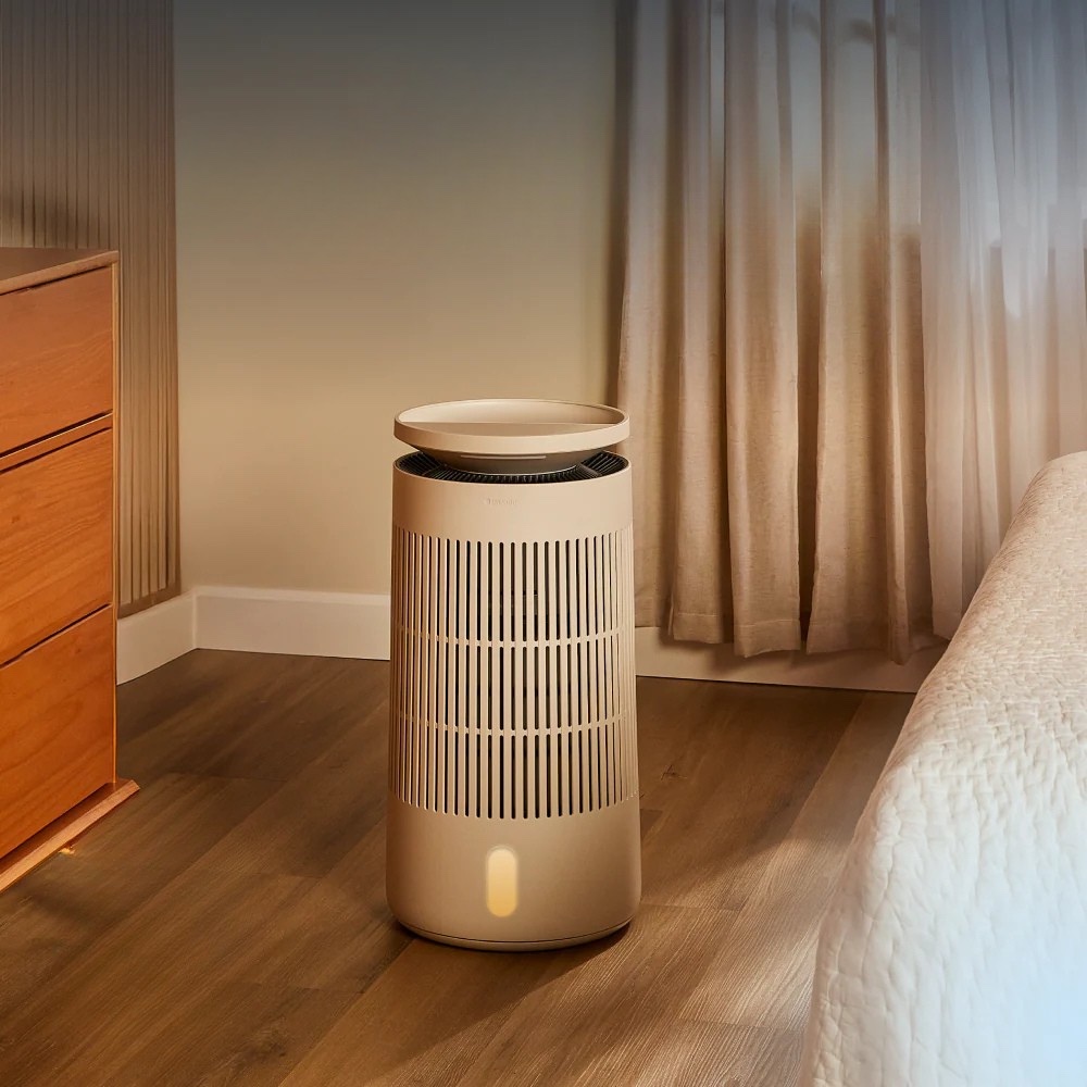 A beige air purifier stands on a wooden floor next to a bed, with a dresser and sheer curtains in the background.