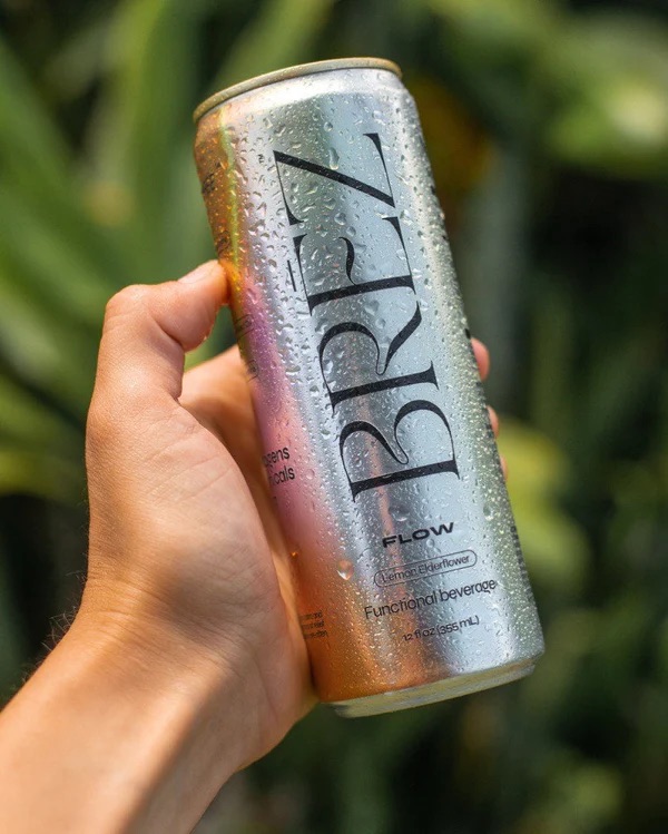 A hand holds a cold, silver can of BREZ Flow functional beverage against a blurred green outdoor background.