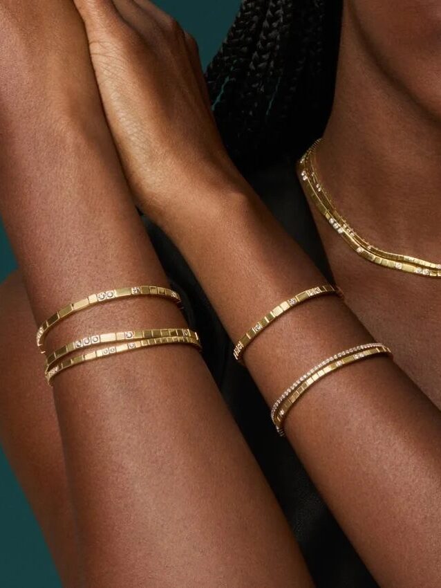 A person with braided hair wears gold bracelets and a gold necklace, with arms crossed over their chest against a dark green background.