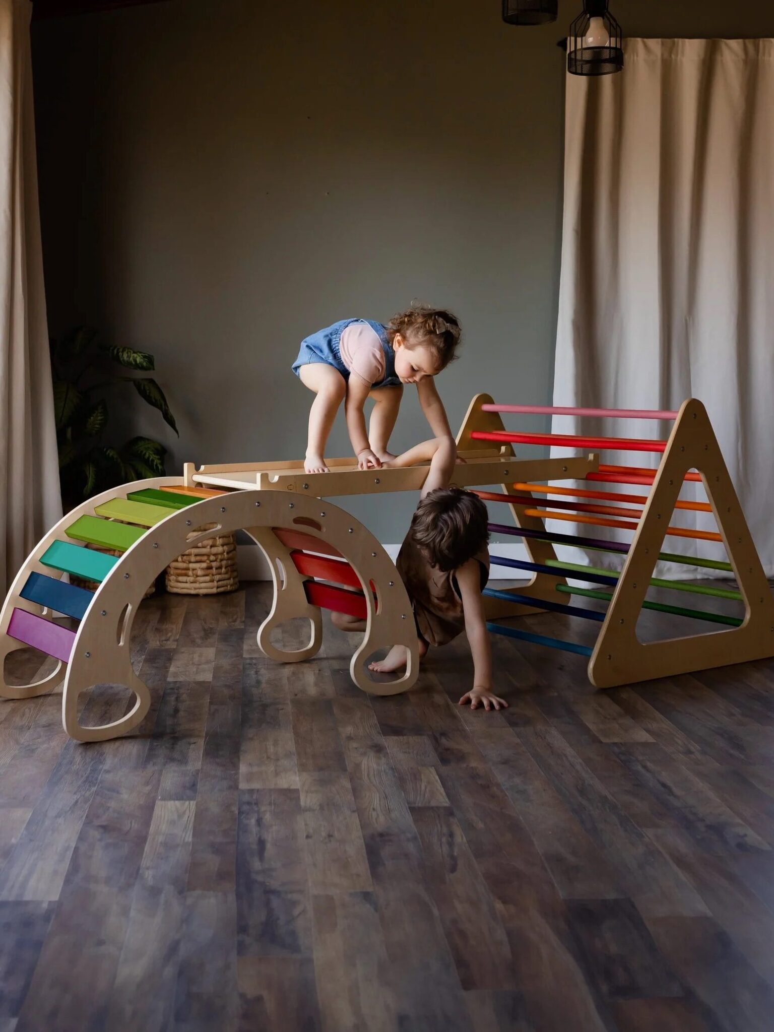 Two children play on a wooden indoor climbing set with arches, ramps, and rainbow-colored bars on a hardwood floor.
