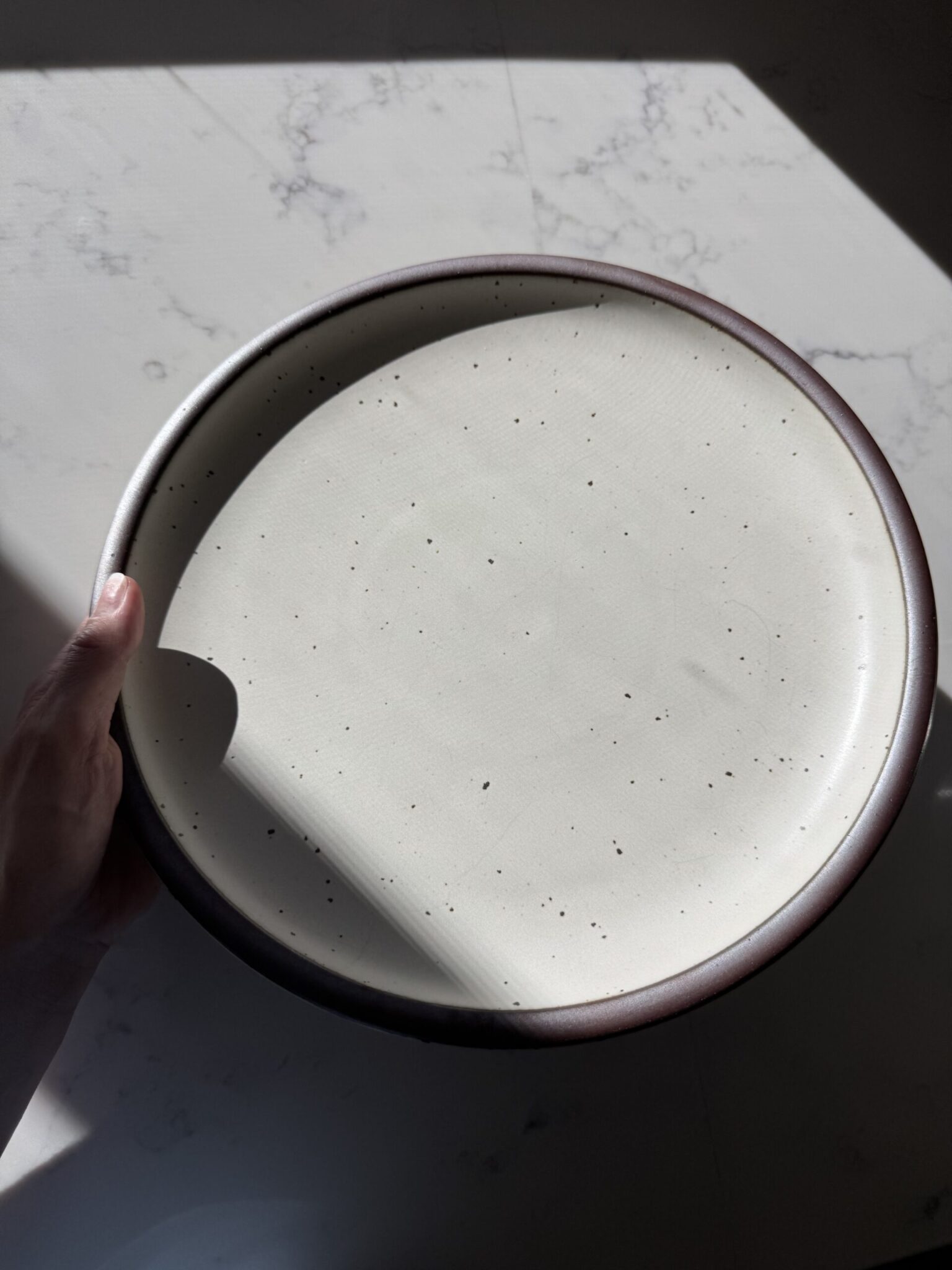 A hand holds a round, speckled ceramic plate in natural light on a light-colored, slightly marbled surface.