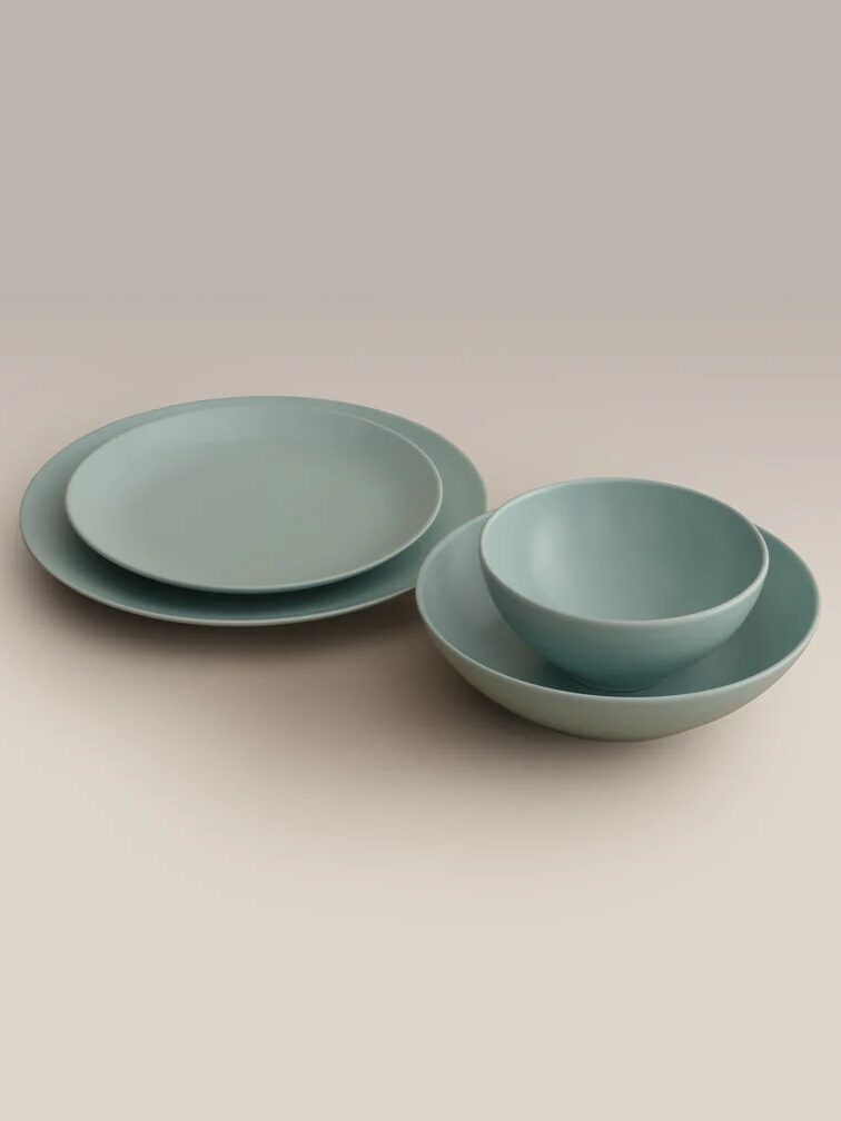 A set of matte light blue dishware including two plates, one bowl, and one shallow bowl, arranged on a neutral background.