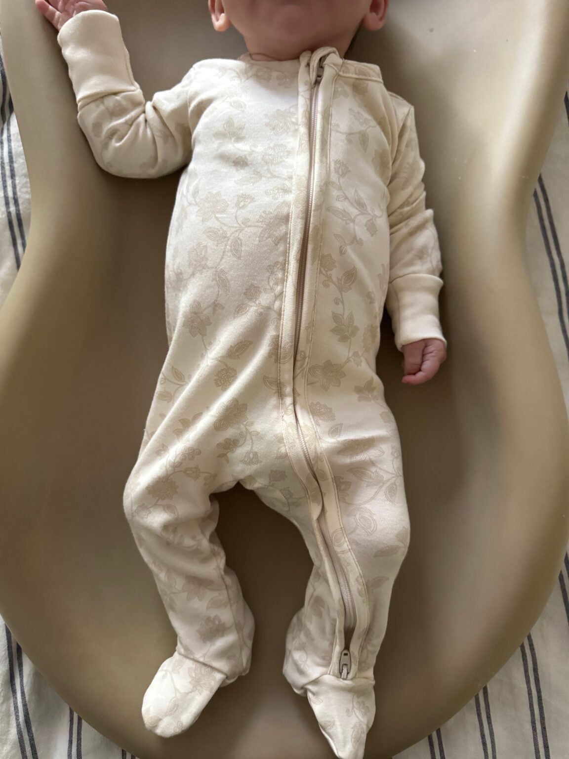 A baby in a cream-colored, floral-patterned zip-up onesie lies on a tan contoured surface with black and white striped fabric underneath.