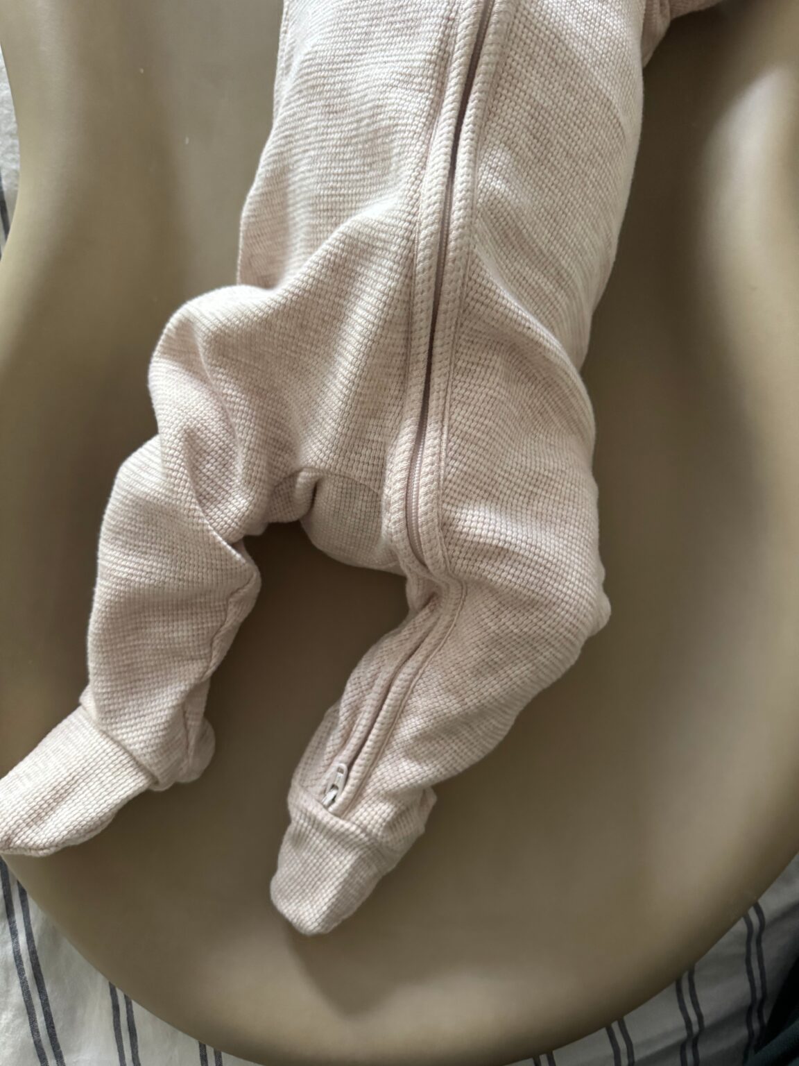 A baby in a textured, light-colored onesie lies face down on a taupe surface, with legs extended and arms out of view.