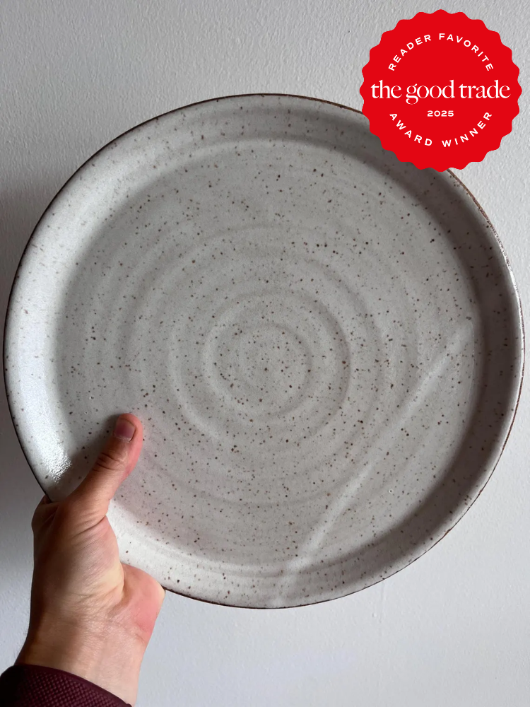 A hand holds a large, round, speckled ceramic plate in front of a white wall. A red badge in the corner reads "The Good Trade 2025 Award Winner, Reader Favorite.