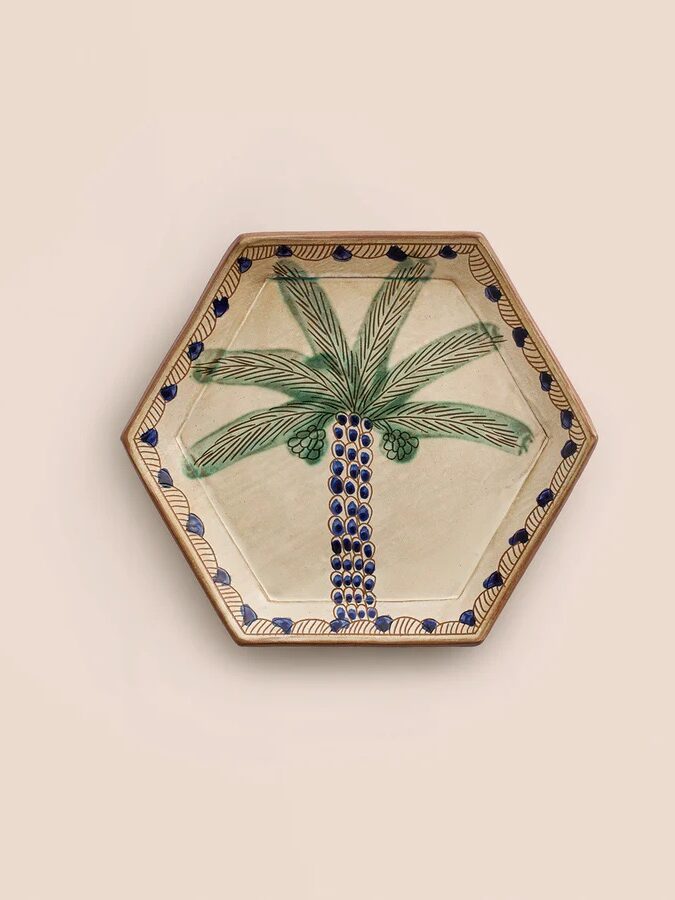 Hexagonal ceramic plate featuring a hand-painted green palm tree with blue fruit and a decorative blue and yellow border on a neutral background.