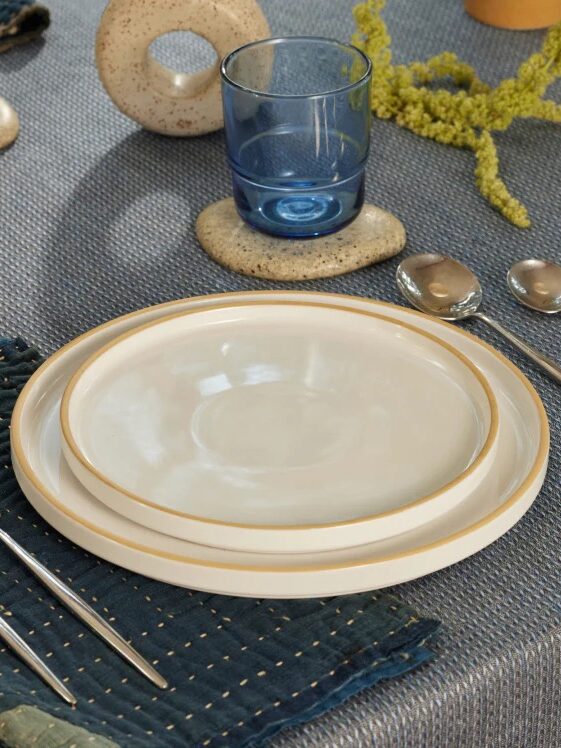 A table set with two stacked white plates, two forks on a dark napkin, two spoons, a blue glass, and decorative items on a textured gray tablecloth.